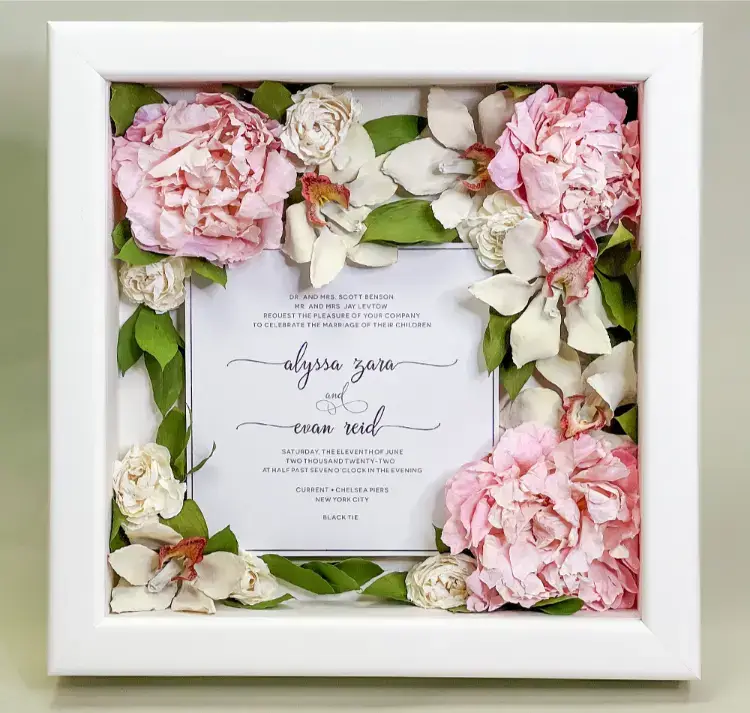 Preserved pink wedding flowers and greenery in a white shadowbox with a wedding invitation.